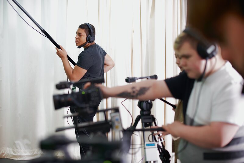 Boom operator in headphones holding a microphone pole beside a camera crew.