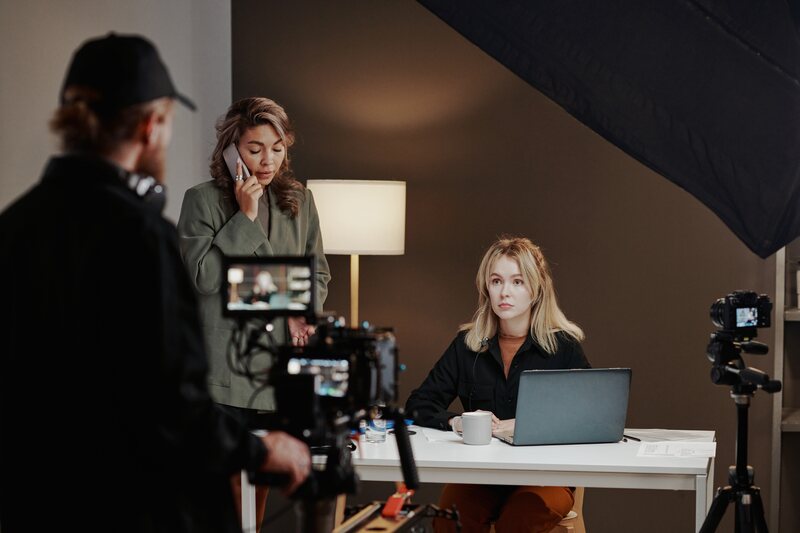 Camera operator filming a woman at a desk under a softbox while a colleague takes a call.