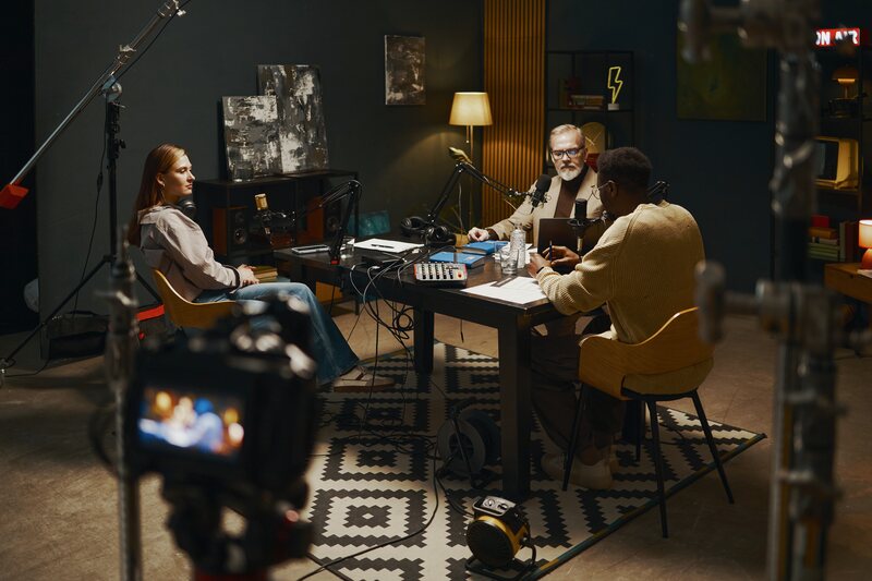 Three people recording a podcast at a table, with a camera and its monitor in the foreground.
